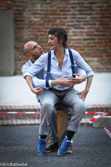 Spectacle - théâtre de rue Deux personnes assises dos à dos, habillées en tenue décontractée avec des bretelles.