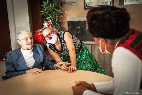 Clowns en maison de retraitre Deux personnes interagissent joyeusement avec une personne âgée autour d'une table.