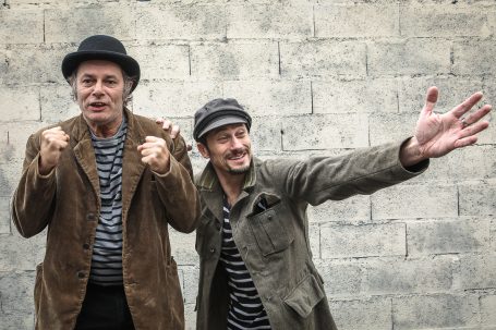 Spectacle - comm cie comédiens Deux hommes en costume rayé, posant joyeusement devant un mur gris.