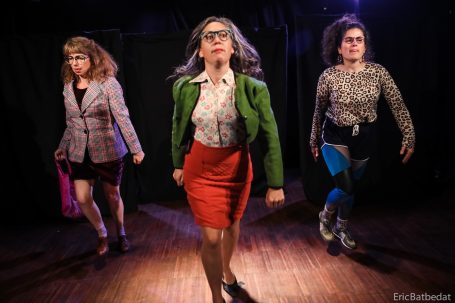 Spectacle - théâtre Trois femmes en tenues variées marchent avec assurance sur scène, éclairées par des projecteurs.