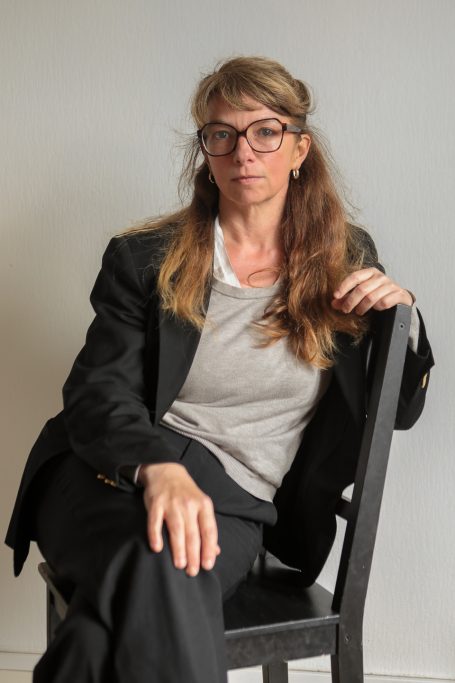 Book comédienne Femme assise sur une chaise, portant des lunettes et un costume noir.