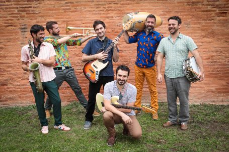 Communication musiciens Groupe de musique composé de six membres, souriants, sous un mur de briques.