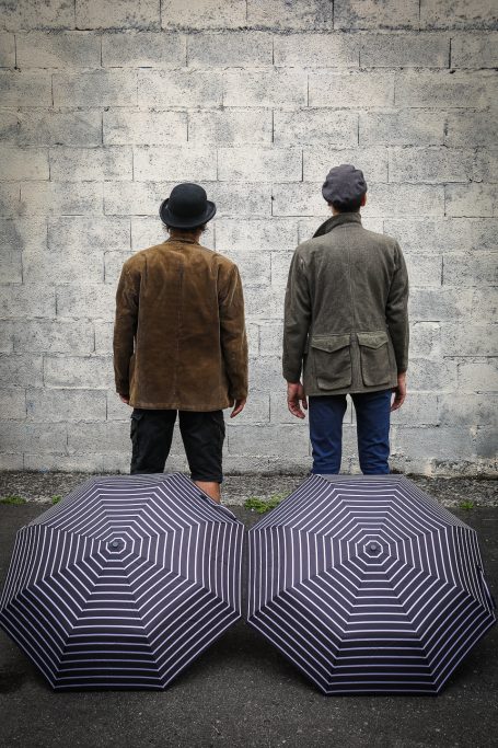 Spectacle - comm cie comédiens Deux hommes de dos, portant des chapeaux, devant un mur, avec deux parapluies rayés à leurs côtés.